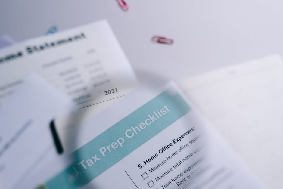 Close-up of a printed tax prep checklist with a light blue header titled 'Tax Prep Checklist' and numbered items related to home office expenses, with a blurred background showing additional sheets of paper and colourful paper clips, all situated on a white surface. The scene suggests preparations for moving or home relocation processes involving packing, organizing, and logistical planning. The checklist is partially visible, and the overall setting indicates an organized approach to managing relocation or moving costs, relevant to house removal services provided by Man and Van Merton as seen on their website page about avoiding hidden removal fees in Merton.