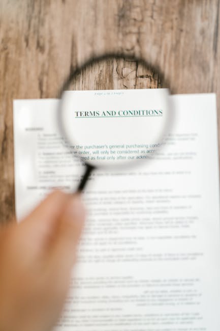 A close-up photograph showing a hand holding a magnifying glass over a printed document titled 'TERMS AND CONDITIONS' on a wooden surface. The magnifying glass enlarges the heading text, making it clearly legible, while the rest of the document appears blurred. The wooden surface beneath the paper is textured, with visible grain and natural imperfections. The image captures a scene related to reviewing contractual or legal documents, which is relevant to home relocation and packing processes involved in removals and furniture transport by companies such as Man and Van Merton, visible through natural contextual connection. The scene emphasizes careful examination of documents in the context of moving services, subtly supporting the topic of transparent pricing and clear terms associated with house removals.