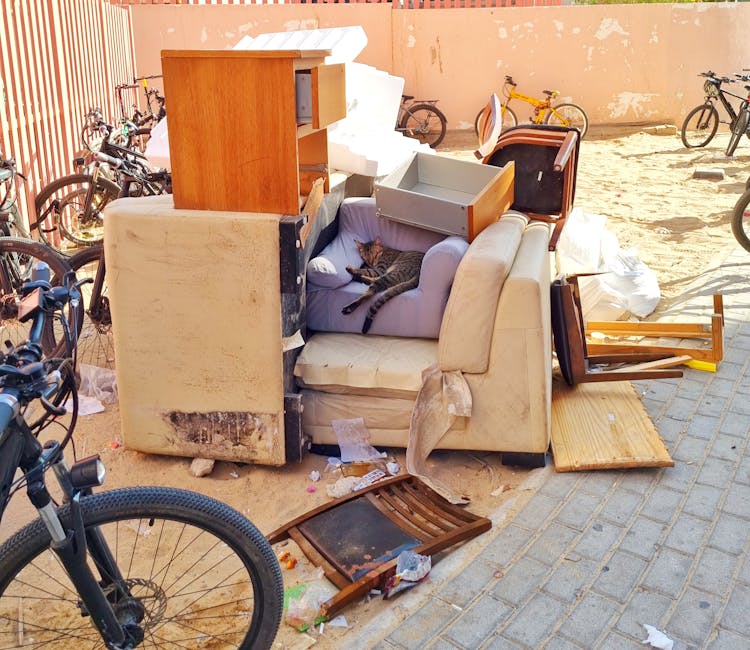 A large pile of household furniture and discarded items outside on a paved sidewalk, arranged for removal. Included are an old, stained beige sofa with visible dirt and damage, partially covered by other furniture and packaging, sitting next to a purple armchair with a cat lying on it. On top of the sofa are wooden furniture pieces, including a small cabinet and a drawer, along with foam packaging materials. Nearby are a wooden dining chair, a small wooden table, and a wooden floor panel, some of which are tipped over or broken. Several bicycles are parked against a rust-coloured wall in the background, with additional bicycles visible behind a chain-link fence. The scene suggests the process of bulky waste removal, possibly as part of a home relocation or clearance, carried out by Man and Van Merton as part of their removals services. Bright sunlight illuminates the scene, highlighting the clutter and the variety of household items prepared for disposal or transport.