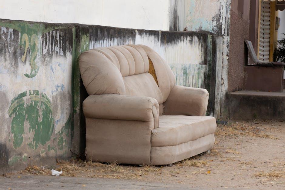 A black and white image showing an ornate vintage sofa with fabric upholstery and wooden armrests, positioned on a sidewalk area. The sofa is located in front of a large, piled wall of crumpled newspapers and cardboard, which fills the background. To the left of the sofa, a person with a blurred, shadowy figure is visible, appearing to either be lifting or positioning the sofa as part of a furniture removal or packing process. The scene is outdoors with natural lighting, and the pavement beneath the sofa is visible. The image reflects a home relocation or furniture transport scenario, with the presence of moving equipment such as straps or trolleys not visible but implied by the context of the scene. The setting underscores the logistical process involved in bulk furniture and packing material disposal or removal, consistent with professionally managed house removals by Man and Van Merton.