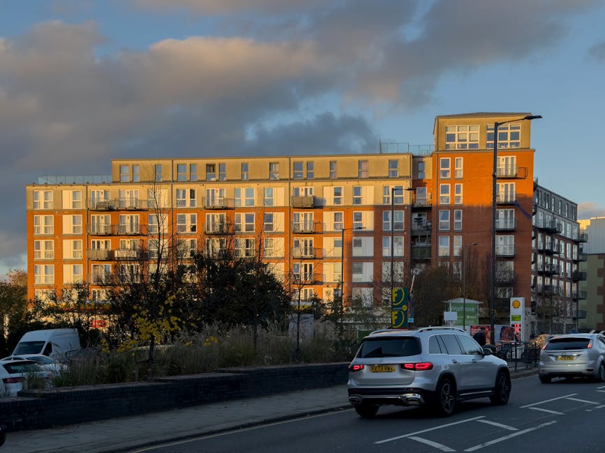 A multi-storey residential building in Colliers Wood with a modern design, featuring numerous balconies with glass railings and large windows. The building is bathed in warm, late-afternoon sunlight, casting a golden hue on the facade. In the foreground, a busy street scene includes several parked and moving vehicles, such as a white van, SUVs, and cars, indicating typical urban traffic. On the pavement, small trees and shrubbery are visible, along with commercial signs for nearby stores or service stations. The scene captures a moment of daily life in Merton, with an emphasis on urban living and transportation context for house removals or furniture transport services provided by companies like Man and Van Merton. The overall setting appears suitable for illustrating home relocation or moving logistics in an urban environment.