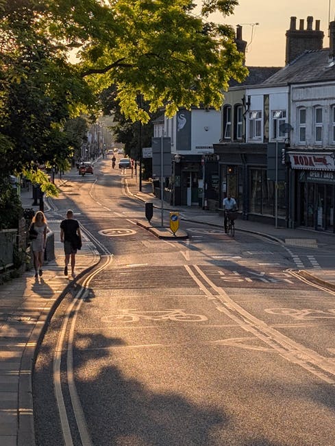 Image of a quiet street in Colliers Wood with a gentle upward slope during late afternoon or early evening, illuminated by warm sunlight filtering through lush green trees lining the sides. Pedestrians, including two women and one man, walk along the pavement on the left side of the road, while a cyclist rides on the right side near a pedestrian crossing. The street features typical UK road markings, including bus lanes and bicycle lanes, with some vehicles visible further down the road. On the right side, there are shops and businesses housed in traditional terraced buildings with dark facades, one of which displays a partial sign. In the background, residential rooftops and chimneys are visible under a clear sky, contributing to a peaceful urban scene. This scene depicts an everyday moment in Merton, relevant to local relocations and transport logistics supported by Man and Van Merton for house removals and furniture transport.