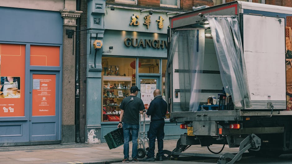Two men are engaged in a home relocation process outside a Chinese restaurant named 'Guang Heung,' situated on a street in Merton. One man, wearing a black cap, jeans, and a dark jacket, is holding a 