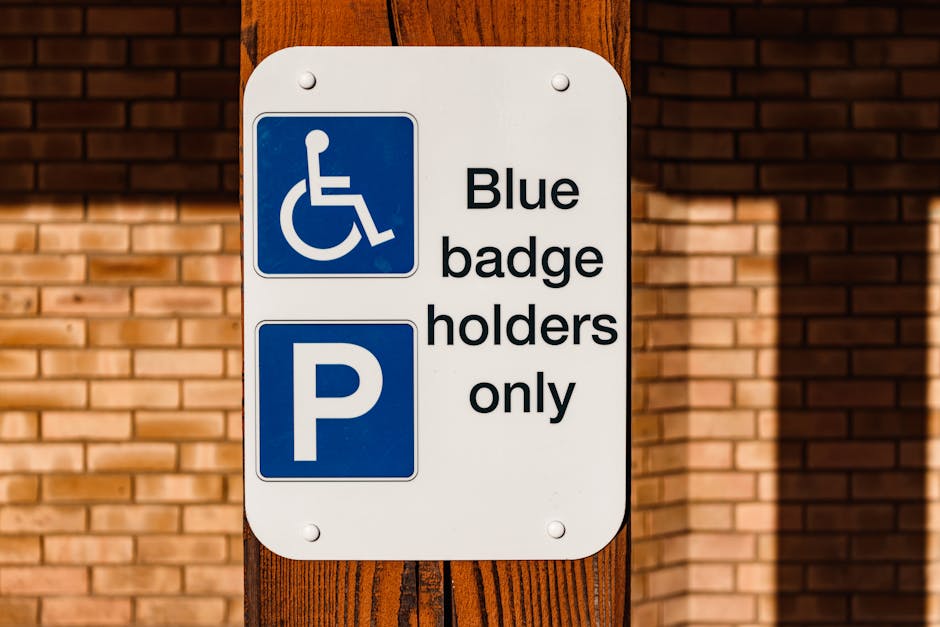 A rectangular white parking permit sign mounted on a wooden post in front of a brick wall. The top section features a blue square with a white wheelchair symbol indicating accessibility, followed by black text that reads 'Blue badge holders only'. Below, a blue square with a white 'P' signifies parking. The sign is outdoors, illuminated by natural daylight, with shadows cast on the wall behind it. The presence of this sign suggests designated parking areas for individuals with blue badges, which is relevant for home relocation and furniture transport services, such as those offered by Man and Van Merton, assisting in moving and logistics within the local area.