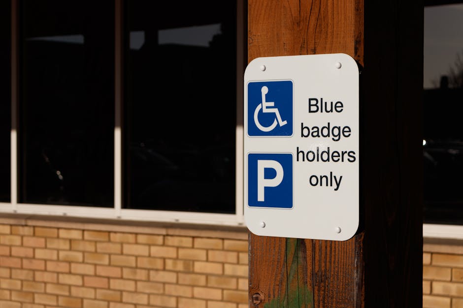 A white rectangular parking sign mounted on a wooden post outside a building with large dark window panels and a brick wall base. The sign features a blue wheelchair symbol at the top, followed by the text 'Blue badge holders only' in black letters. Below, there is a blue square with a white letter 'P,' indicating parking reserved for blue badge holders. The sign's placement suggests designated accessible parking, which may be relevant for a home relocation or removal services context, such as ensuring accessible vehicle parking for furniture transport or loading operations. The background shows a reflective glass window and part of the brick wall, with natural lighting illuminating the scene. Man and Van Merton, a removal company, may use such parking permits to facilitate smooth loading and unloading during house removals, relocations, and furniture transport tasks.