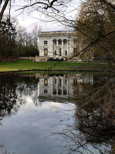 A large, white, neoclassical-style house with tall columns and decorative balcony railings is situated behind a small pond, reflecting the building and surrounding leafless trees in the water. The house is set on a well-maintained grassy area, and an open door is visible on the front of the building. Part of an ongoing home relocation process is suggested by the presence of cardboard boxes, packing materials, and a moving blanket on the pavement near the house’s entrance. A green moving trolley with wheels and straps is positioned nearby, ready for transporting furniture or boxes. The scene is outdoors during overcast weather with natural daylight, and the house appears to be part of a relocation or furniture transport operation managed by Man and Van Merton, as indicated by the context of house removals and moving services in the area.