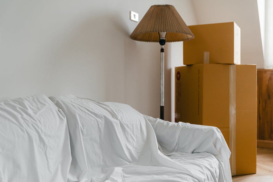 Inside a room showing a white sofa partially covered with a white sheet, positioned against a plain beige wall. Next to the sofa is a tall floor lamp with a brown conical lampshade. Behind the lamp, there are three cardboard boxes stacked in a corner, with the top box slightly offset. The boxes are sealed with clear packing tape and vary in size. The room has wooden flooring and natural light coming through a nearby window, creating a well-lit environment. The scene suggests preparations for a home relocation or packing process, with furniture and household items ready for transport by a professional removals service such as Man and Van Merton.