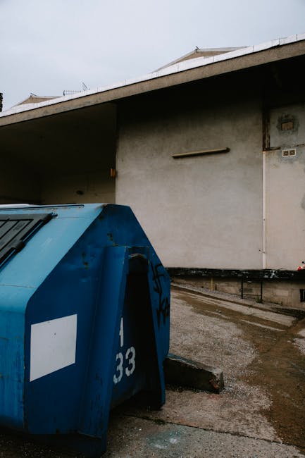 A blue metal refuse bin with a white label and the number 33 on its side is positioned on a concrete driveway outside a building, next to a small concrete curb. The bin is situated near the edge of the pavement, with the rear facing towards the building's exterior wall, which is plain and painted in light grey. The area appears to be part of a residential or commercial property, with the driveway leading to the building's entrance. The scene is outdoors under overcast lighting conditions. This setting potentially shows part of a home relocation or clearing process involving removal or disposal of waste materials, consistent with the services offered by Man and Van Merton for house removals and moving logistics.