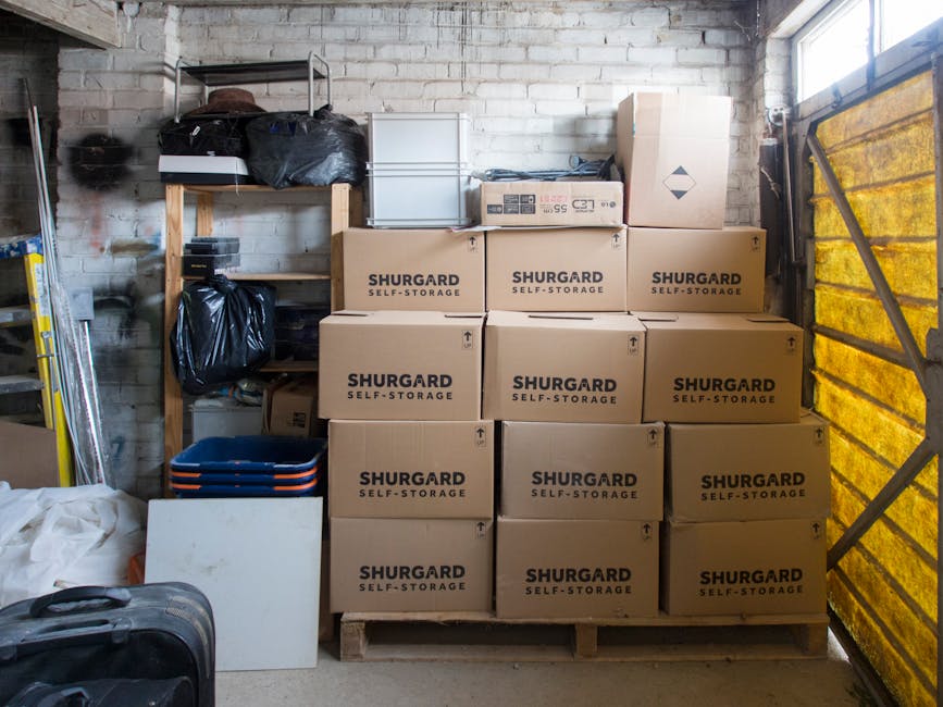 Inside a storage space associated with a house relocation, a wooden shelving unit is positioned against a white brick wall, with various black plastic bags and small boxes placed on the upper shelf. In front of the shelving, a stack of brown cardboard moving boxes labeled 'SHURGARD SELF-STORAGE' is arranged on a wooden palette, forming a compact storage block. To the left, a white panel leans against the boxes, and a set of blue and orange plastic stacking crates is visible beneath part of the shelving. To the right, a partially visible yellow-tinted transparent sliding door or window panel provides natural light, casting illumination across the area. The scene captures the interim storage of packing materials, boxes, and personal items, reflecting the practical short-term solutions often used during a house move when access to organized storage space is limited, with the environment ready for furniture transport and home relocation activities as managed by Man and Van Merton.