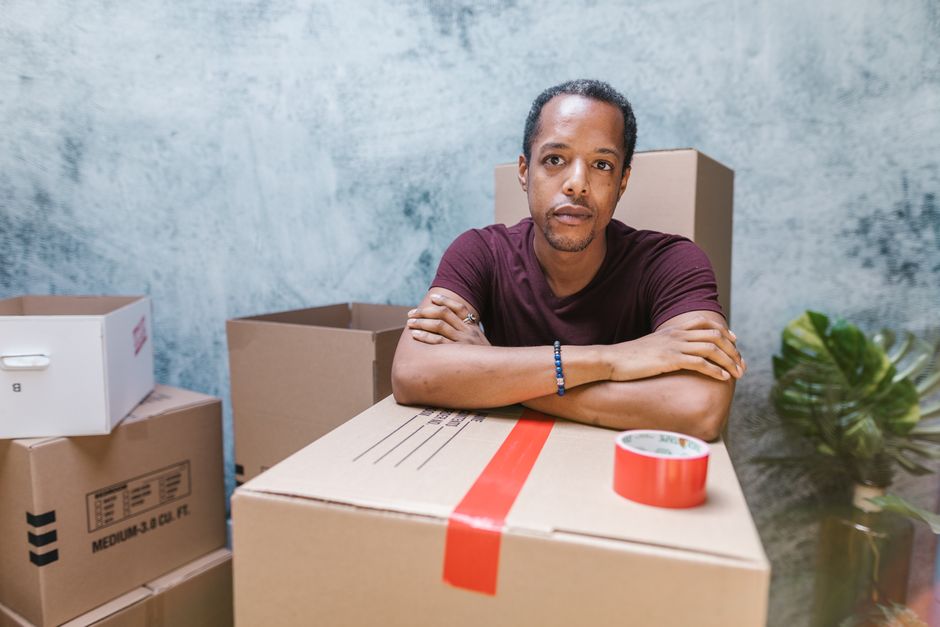 A man with short dark hair, wearing a maroon T-shirt and a blue beaded bracelet, sits behind a large cardboard box with red packing tape, inside a room with a textured grey wall. Surrounding him are several packed cardboard boxes, some labeled, and a roll of red packing tape on the table. To the right, a green houseplant is partially visible on a dark surface. The scene depicts a home relocation or furniture transport process, with the man appearing to oversee or await the next phase of packing or moving, characteristic of the packing and moving activities carried out by Man and Van Merton during a short-term storage after a failed move at the property.