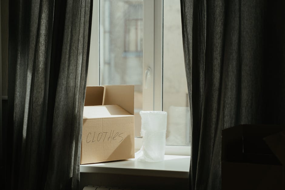 Inside a storage space associated with a house relocation, a wooden shelving unit is positioned against a white brick wall, with various black plastic bags and small boxes placed on the upper shelf. In front of the shelving, a stack of brown cardboard moving boxes labeled 'SHURGARD SELF-STORAGE' is arranged on a wooden palette, forming a compact storage block. To the left, a white panel leans against the boxes, and a set of blue and orange plastic stacking crates is visible beneath part of the shelving. To the right, a partially visible yellow-tinted transparent sliding door or window panel provides natural light, casting illumination across the area. The scene captures the interim storage of packing materials, boxes, and personal items, reflecting the practical short-term solutions often used during a house move when access to organized storage space is limited, with the environment ready for furniture transport and home relocation activities as managed by Man and Van Merton.