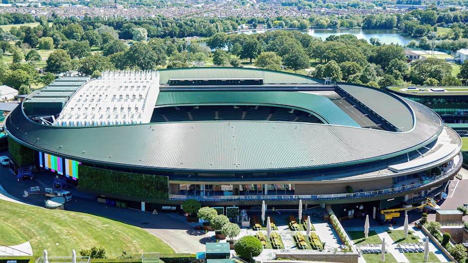 Aerial view of a large, modern sports stadium with a curved, green metal roof covering the seating areas and an open central section for the pitch. Surrounding the stadium are well-maintained green lawns, small trees, and outdoor seating areas with yellow and white tables and umbrellas. The stadium is located in a lush, green environment with a mix of trees, open fields, and a body of water visible in the background, suggesting a peaceful, suburban setting. Near the entrance, vehicles including a van and a small truck are parked, indicating an active loading or moving process. The lighting is bright and clear, highlighting the structural details of the stadium, which is associated with sports events and large-scale gatherings, relevant for sports venue management or event logistics, similar to professional removals or transport planning for large venues.