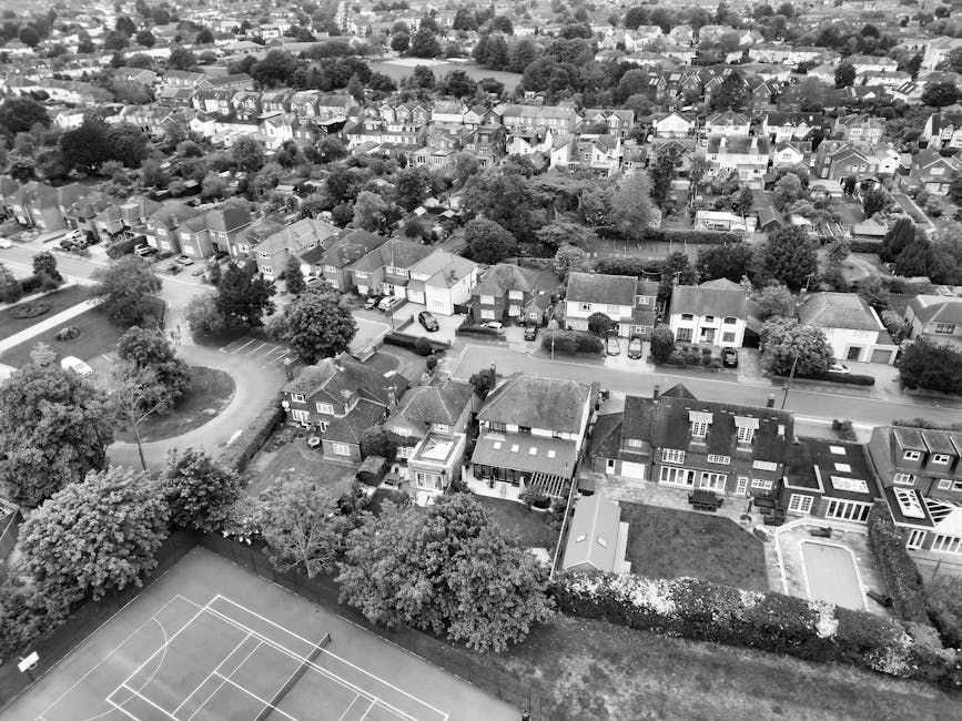 An aerial black-and-white photograph of a residential neighbourhood showing a row of detached and semi-detached houses with front gardens, some featuring outdoor seating areas and driveways with parked cars. Several trees line the street, providing greenery among the houses. In the lower left corner, there is a sports court, possibly for tennis or basketball, with visible white court lines. In the foreground, part of a property boundary fence and rooftops are visible, indicating the view is taken from a drone or high vantage point. The scene captures a quiet, suburban area that could be a typical setting for house removals or home relocation services, illustrating the environment in which Man and Van Merton operates for house removals, furniture transport, and packing and moving projects in Wimbledon Village (SW19).